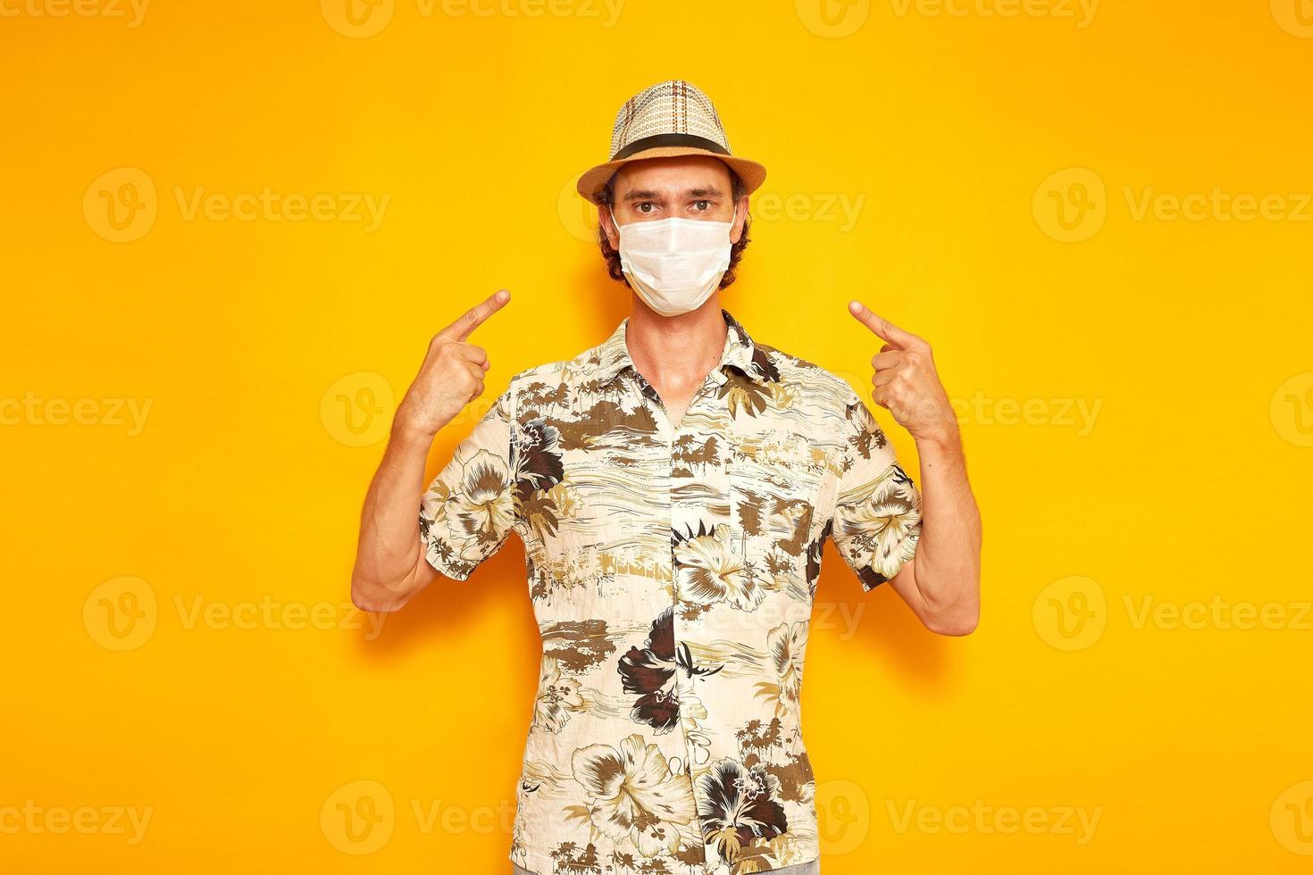 male tourist traveler points with his index fingers at a medical mask on his face. isolated on a yellow background with space for text. tconcept of people, vacation, protection from virus covid-19 photo