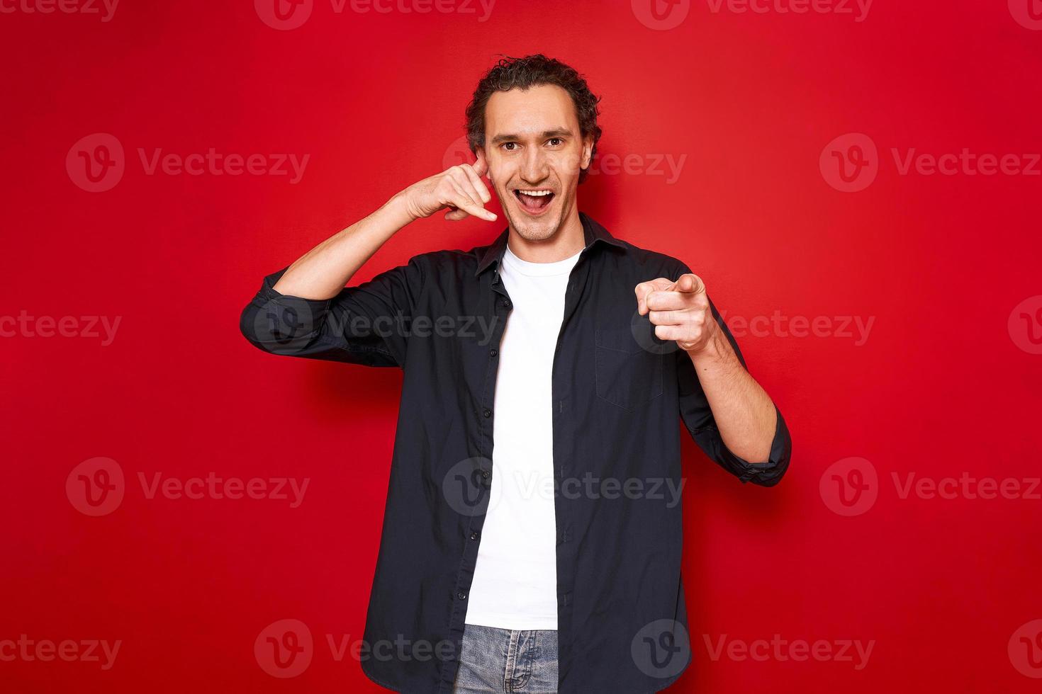 call me hand sign. portrait of young smiling man pointing index finger in front of him depicts telephone receiver in his hand. isolated on red studio background. concept - love, romance, communication photo