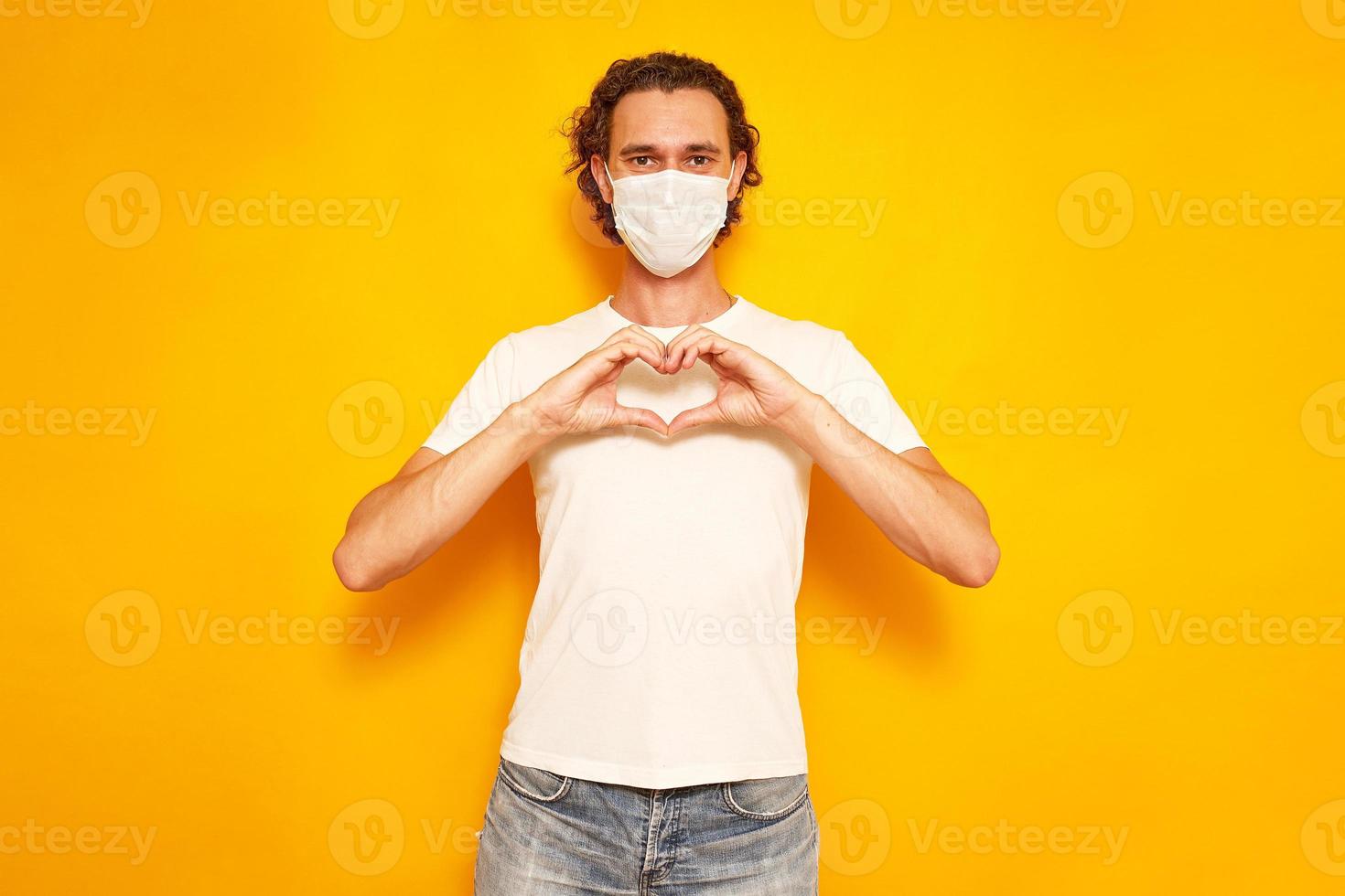 portrait of young man in medical mask folded his hands in form of heart in front of him on his chest. isolated on yellow background. space for text. concept of love, gratitude, sympathy, valentine day photo