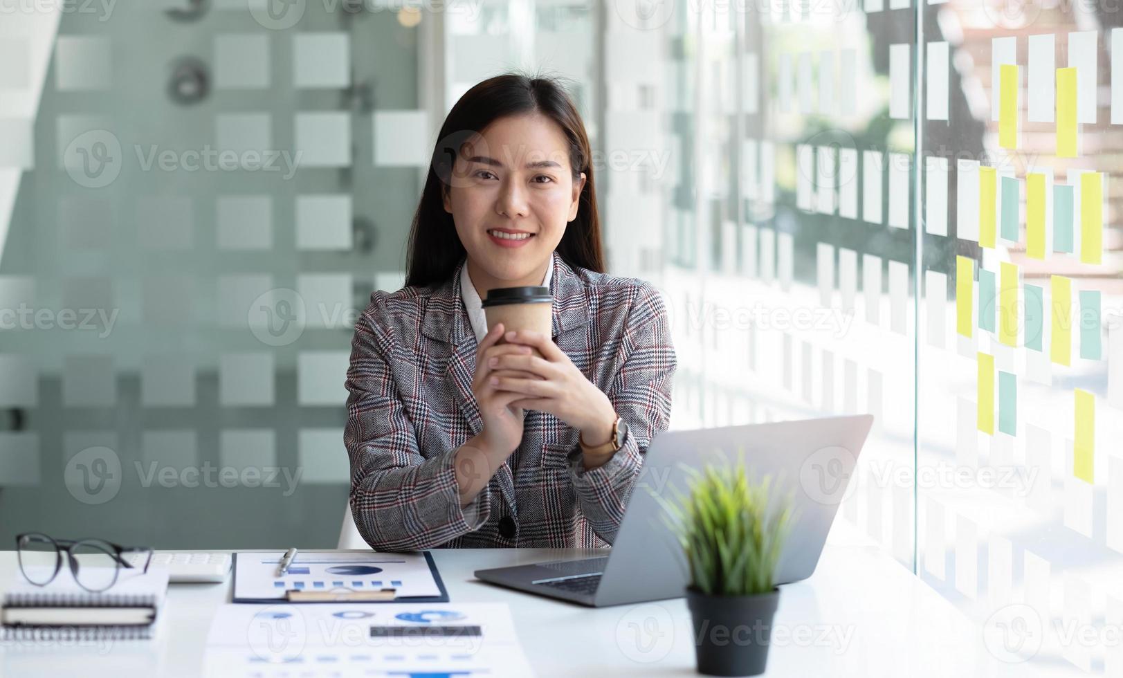 encantadora mujer de negocios asiática parada con una taza de café mirando la cámara en la oficina. foto
