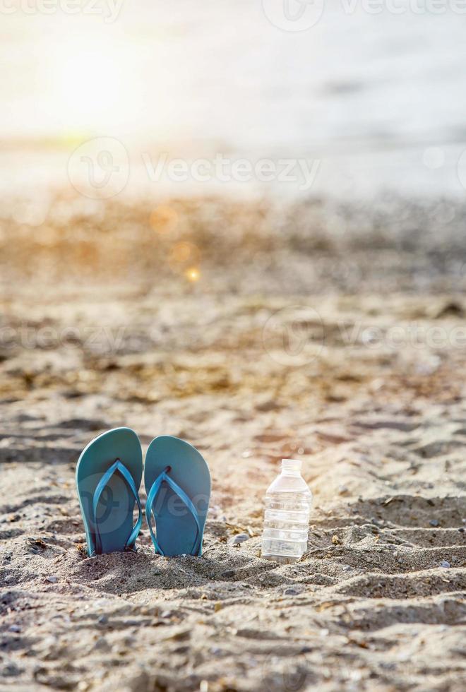 Flip flops on beach sand with water in plastic bottle and sunbeam photo