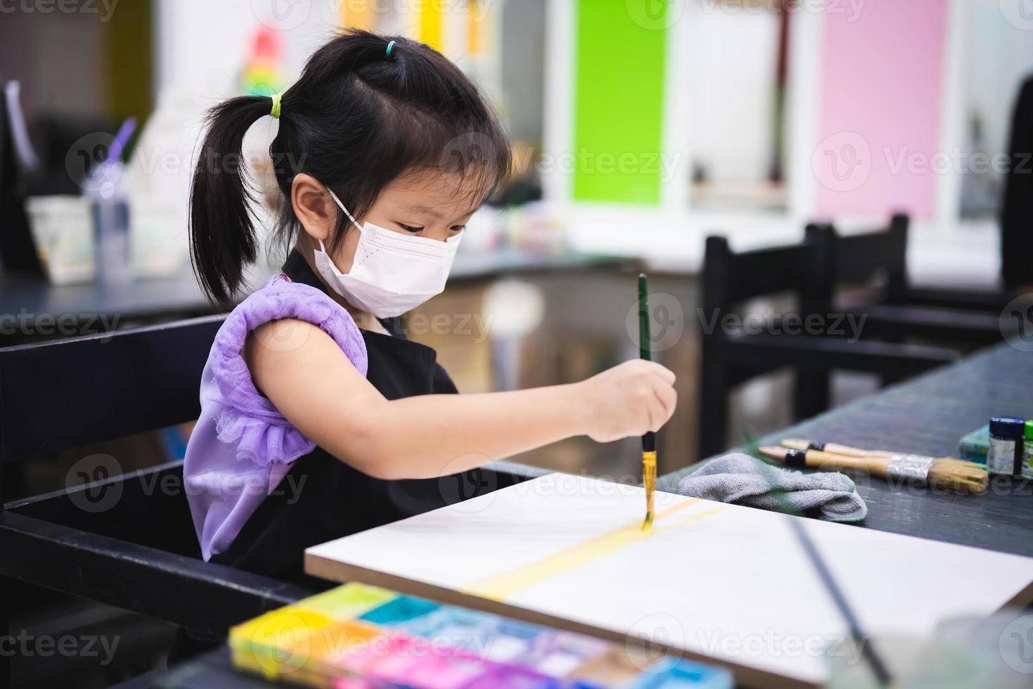 Portrait cute kid girl wearing white face mask. Child learn to paint in classroom. Children holds brush to paint on canvas. Kid 5  year old does art. photo