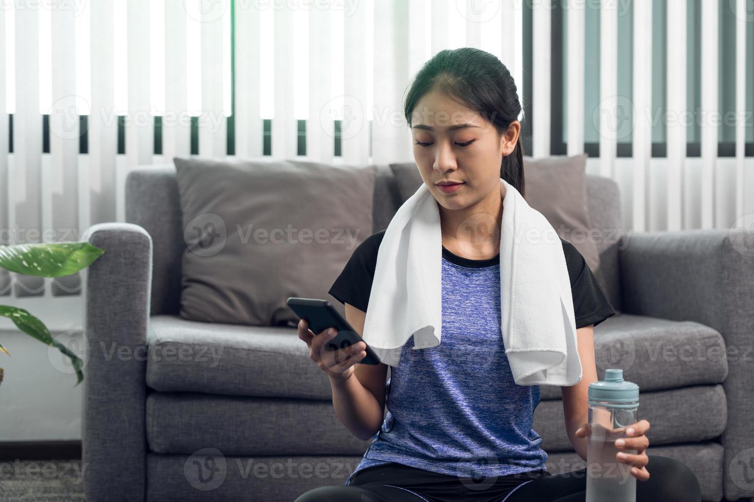 Asian woman playing smartphone at home during break from yoga exercise and drinking fresh water. photo