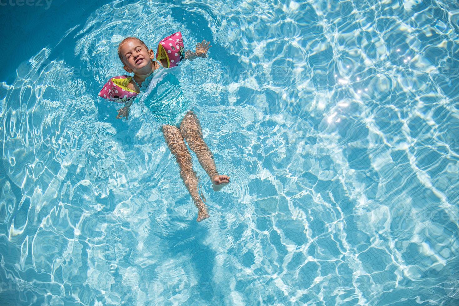 linda niña pequeña nadando en una piscina turquesa con brazos inflables, ayuda para apoyar, vista superior. foto