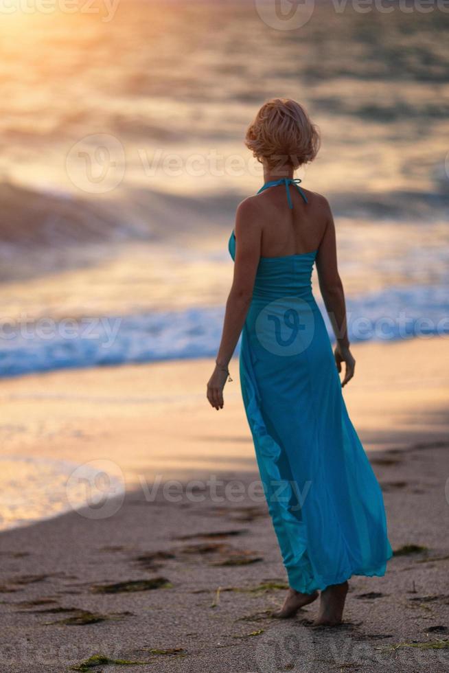 A young slender girl stands on the beach at sunset in a blue long dress, a beautiful figure photo