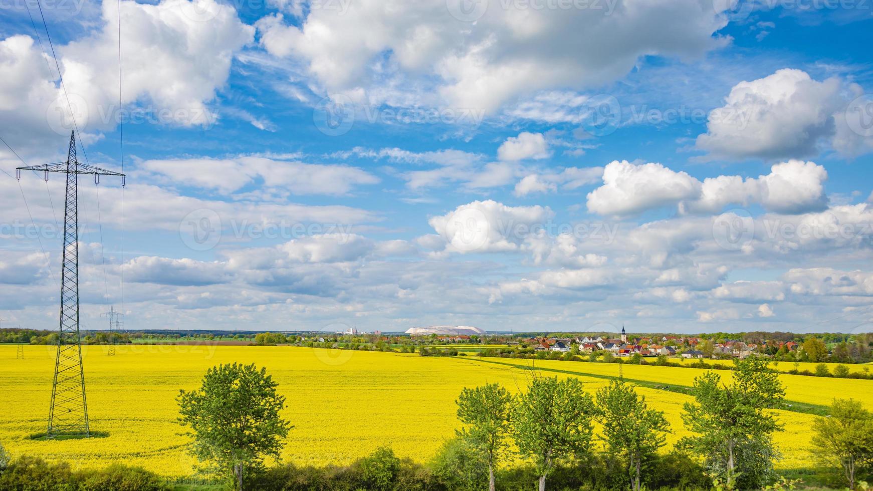 Panoramic view over beautiful farm countryside landscape with rapeseed yellow at blossom field, with a village at horizon in Germany, Spring, and high voltage power lines at blue sky and sunny day. photo