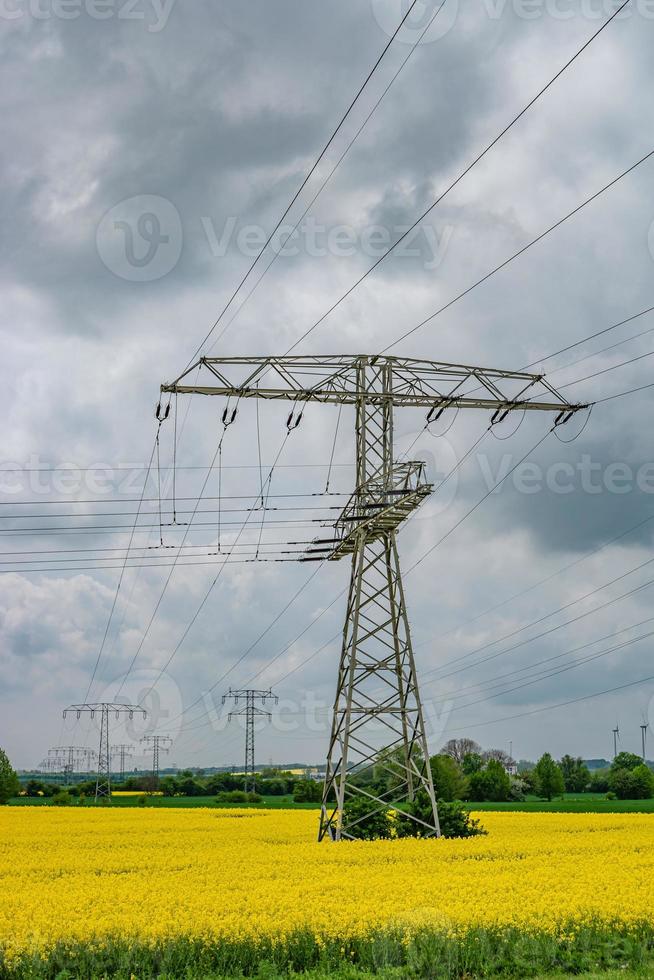 Beautiful farm landscape with yellow rapeseed at blossom field, wind turbines to produce green energy and high voltage power lines in Germany, at Spring and dramatic rainy sky. photo