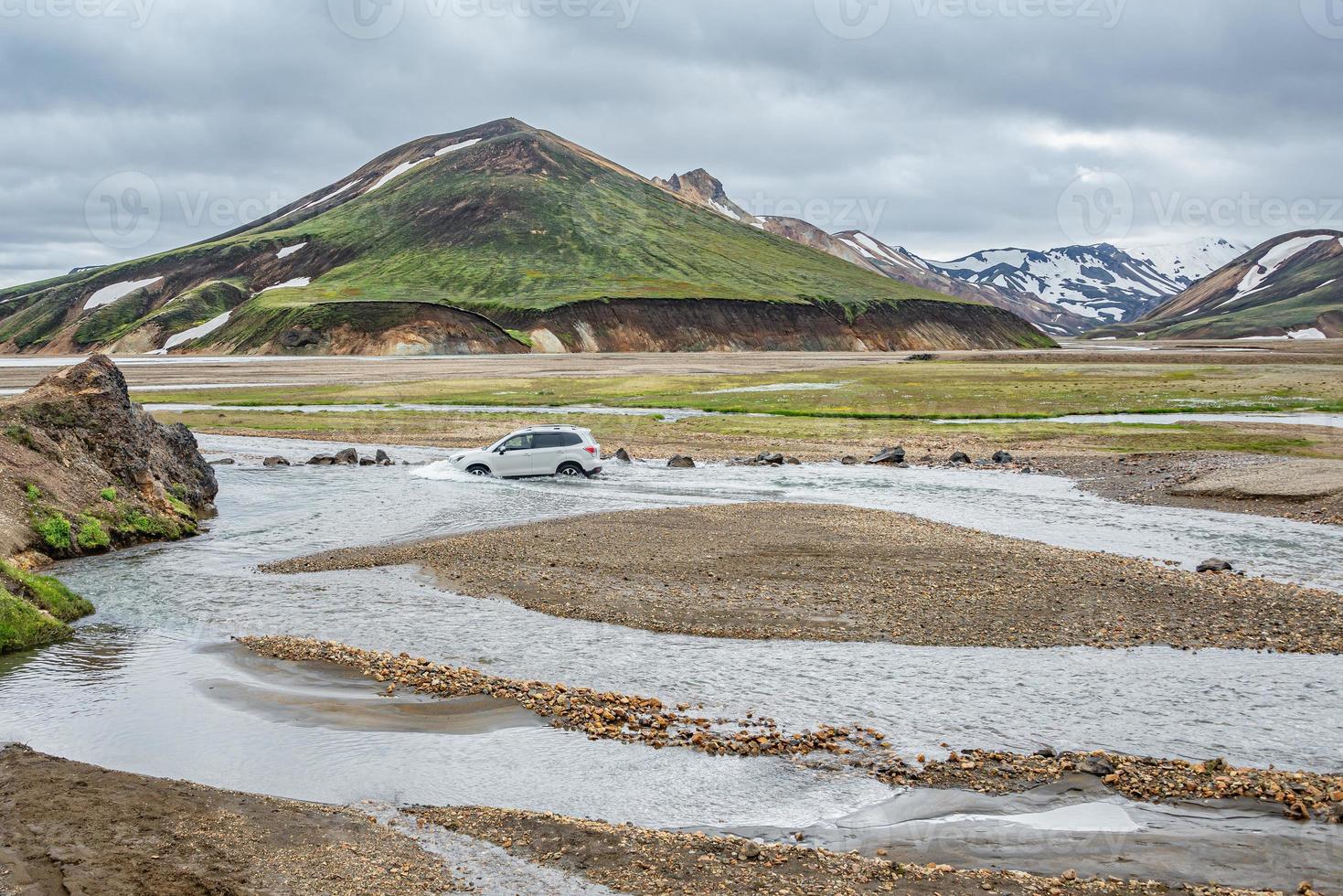 A car is fording a mountain creek at surreal magic Icelandic landscape of colorful rainbow volcanic Landmannalaugar mountains, near a biggest camping site there with tourists and hikers, Iceland photo
