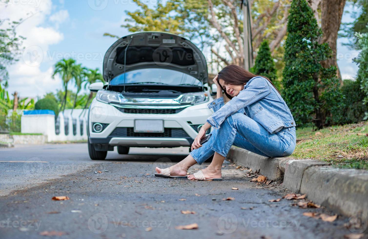 Asian woman sitting beside car after a car breakdown on street. Concept of vehicle engine problem or accident and emergency help from Professional mechanic photo
