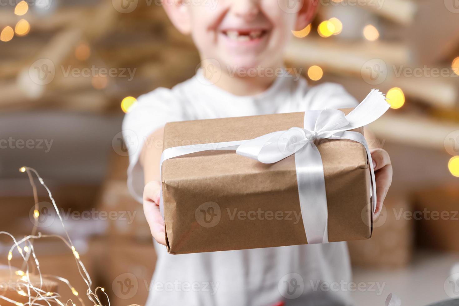 simple gift in kraft paper packaging tied with a white ribbon in the hands of a child boy in a t shirt. baby holding gift box on christmas background. concept of new year, mothers day, Valentines day photo