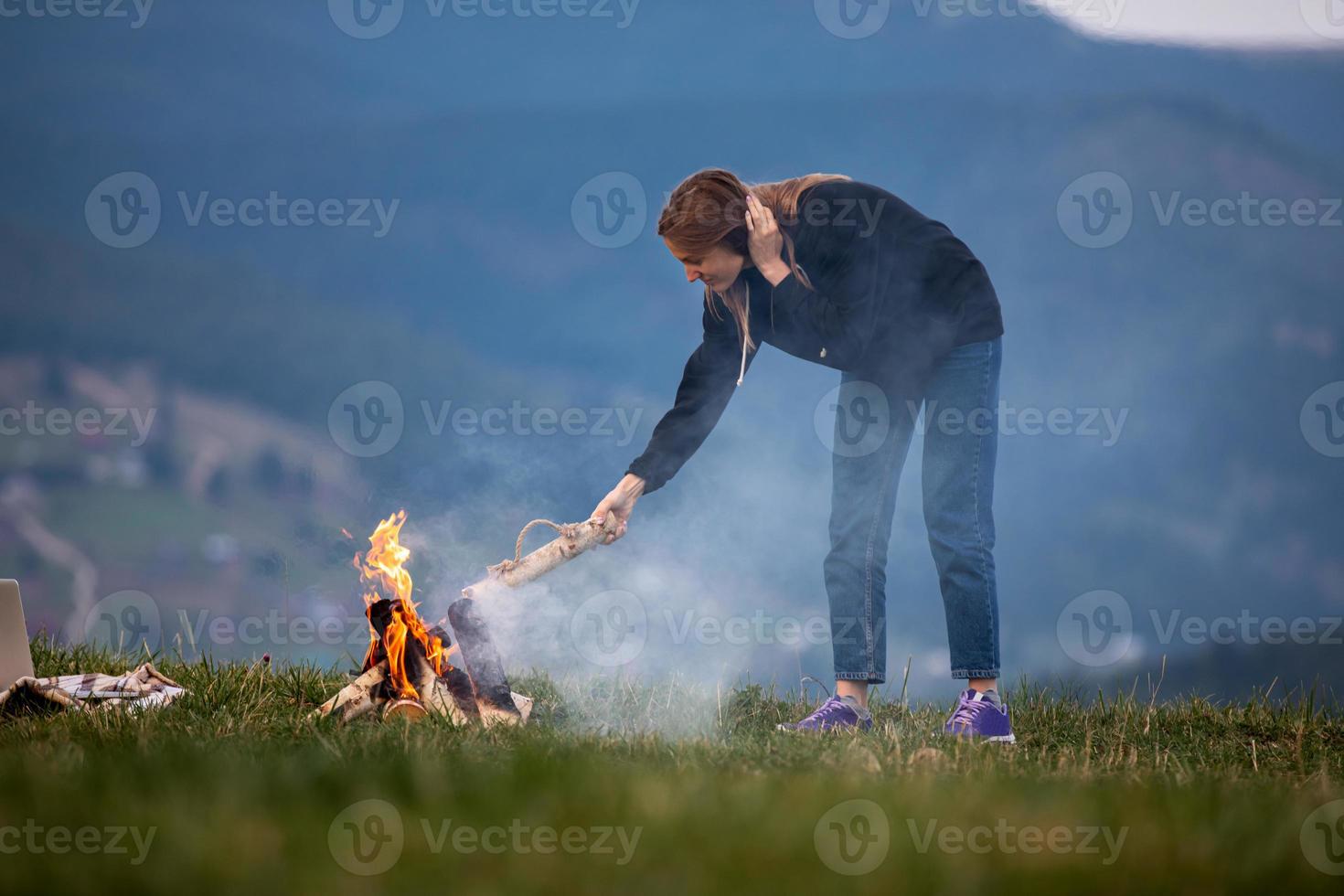 young woman freelancer throws firewood into a fire in the mountains in the evening. tourist girl resting and working outdoor photo