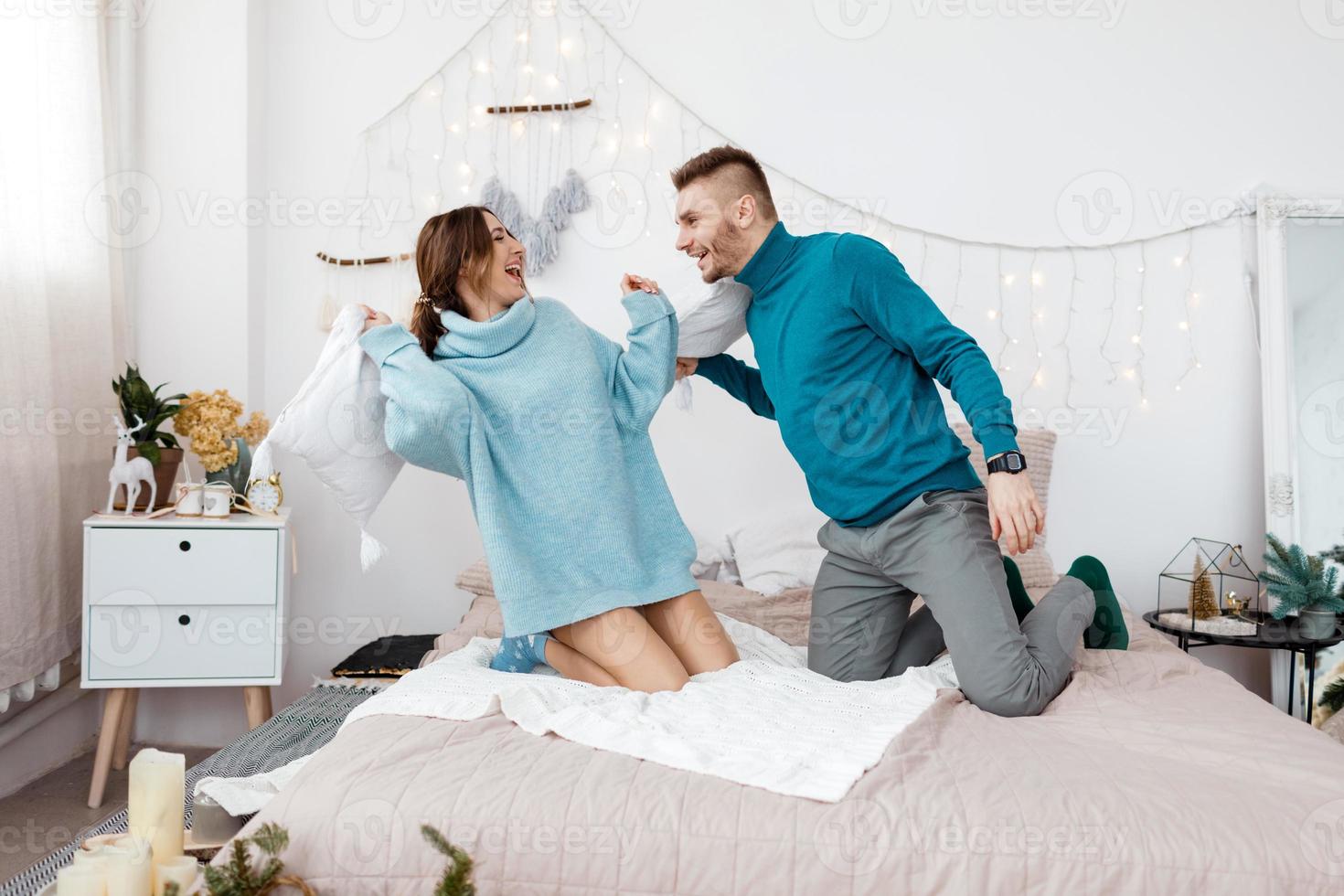 feliz pareja amorosa con estilo teniendo una pelea de almohadas en la cama. joven y mujer esperando bebé para navidad. enfoque selectivo foto