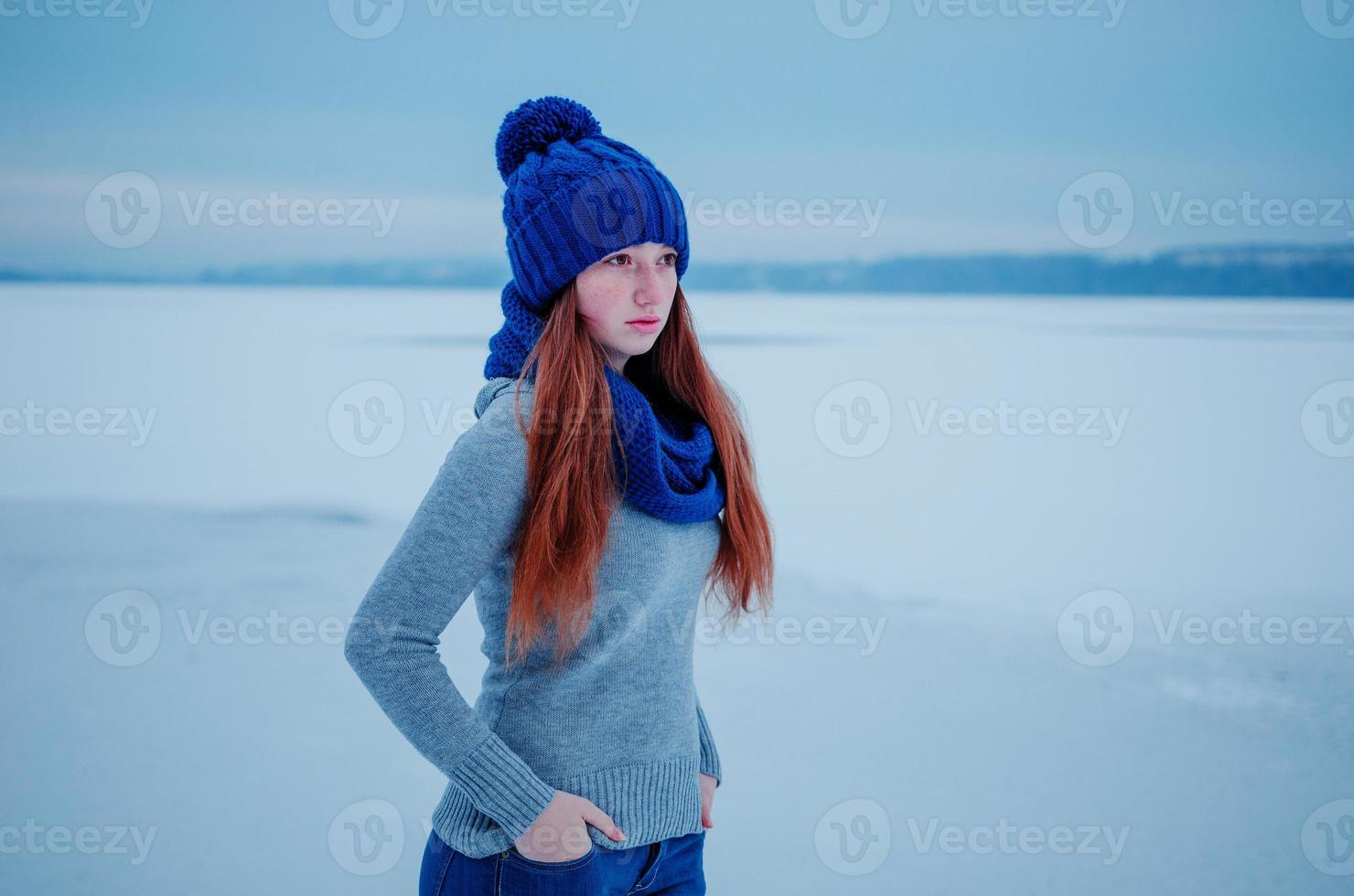 retrato de una joven pelirroja con pecas con sombrero de lana de punto azul y bufanda en el hielo de fondo del día de invierno. foto