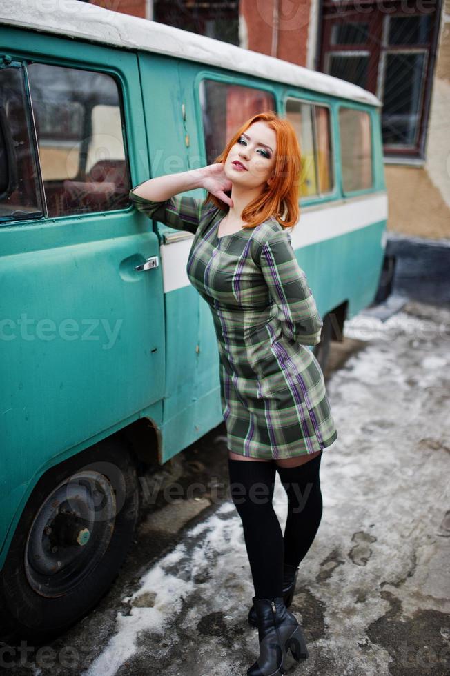 Young red haired girl posed on checkered dress background old retro cyan minivan. photo