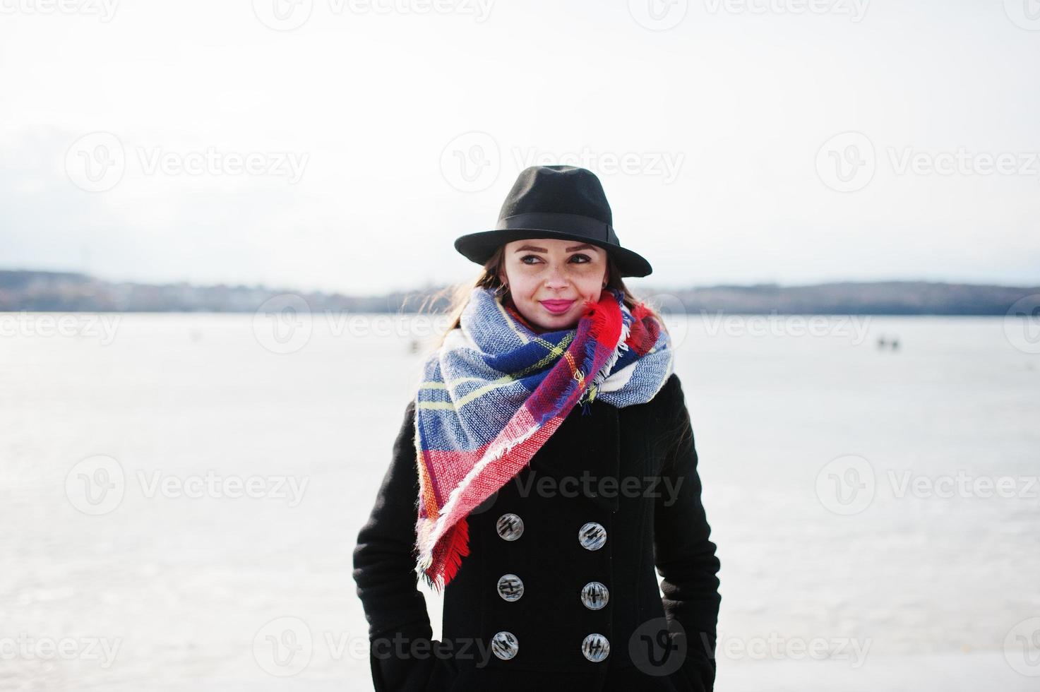 chica joven casual divertida en abrigo negro, bufanda y sombrero contra el río congelado en el clima soleado de invierno. foto