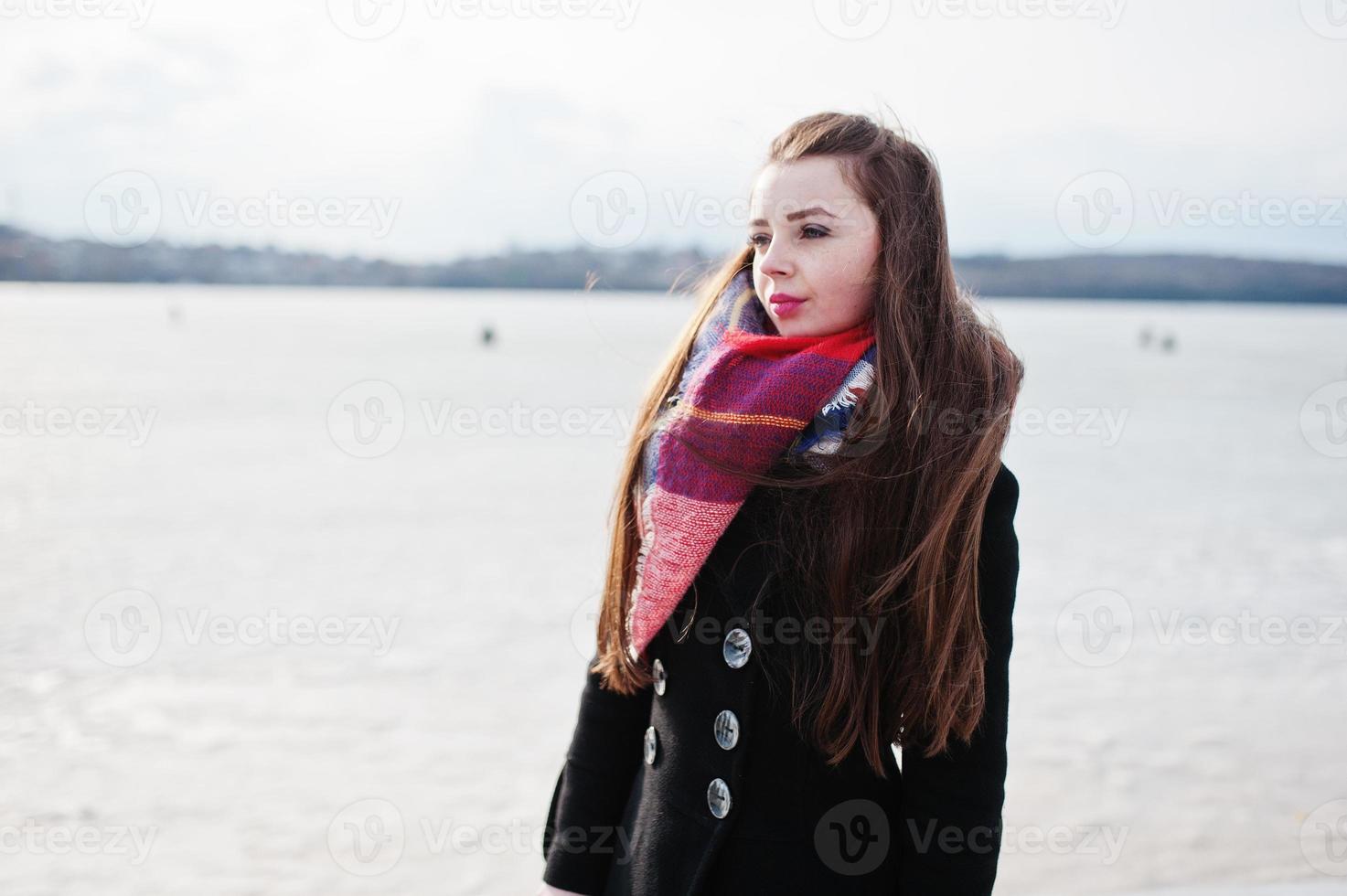 chica joven casual en abrigo negro, bufanda y sombrero contra el río congelado en el clima soleado de invierno. foto