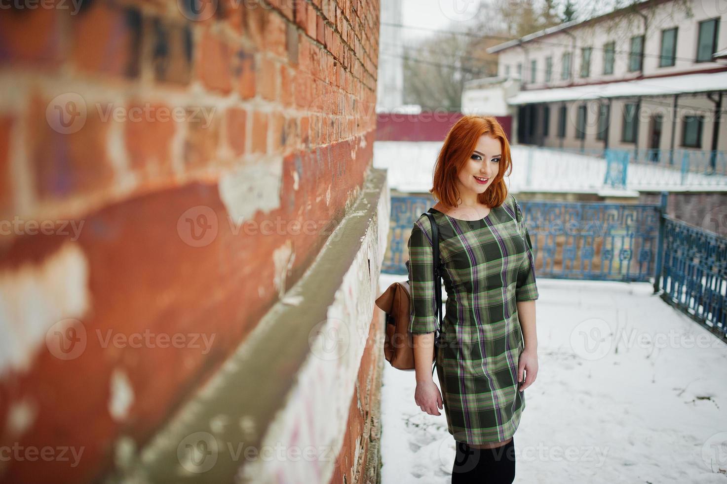 An outdoor portrait of a young pretty girl with red hair wearing checkered dress with girly backpacks standing on the brick wall background in winter day. photo