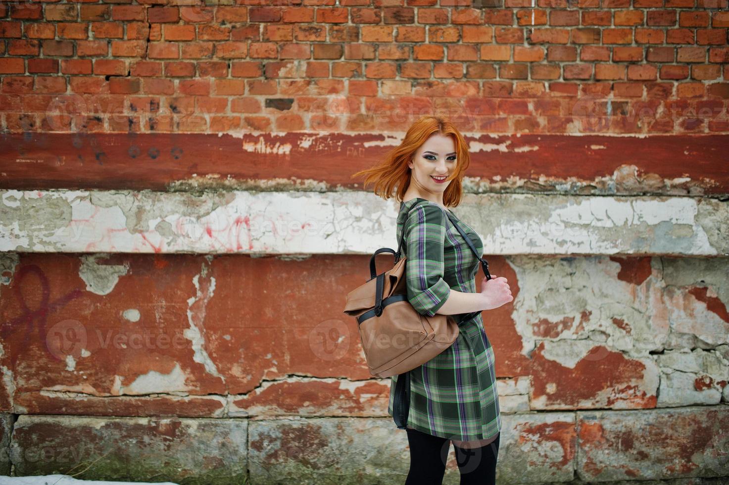 An outdoor portrait of a young pretty girl with red hair wearing checkered dress with girly backpacks standing on the brick wall background. photo