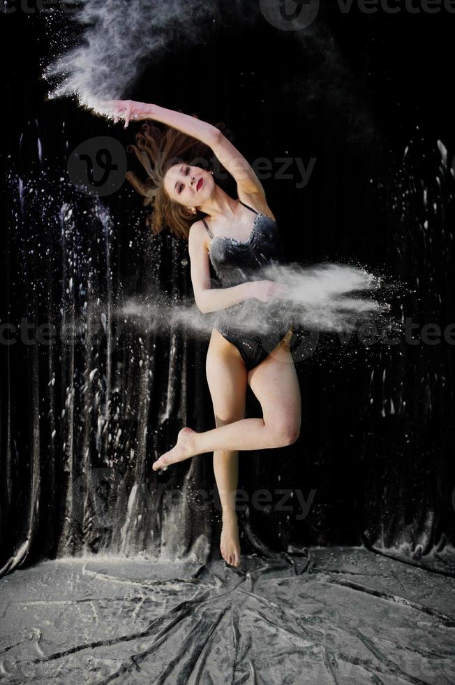 Girl dancer jumping and dancing in the white dust with flour on a black background. Studio shot of woman dancing with flour. photo