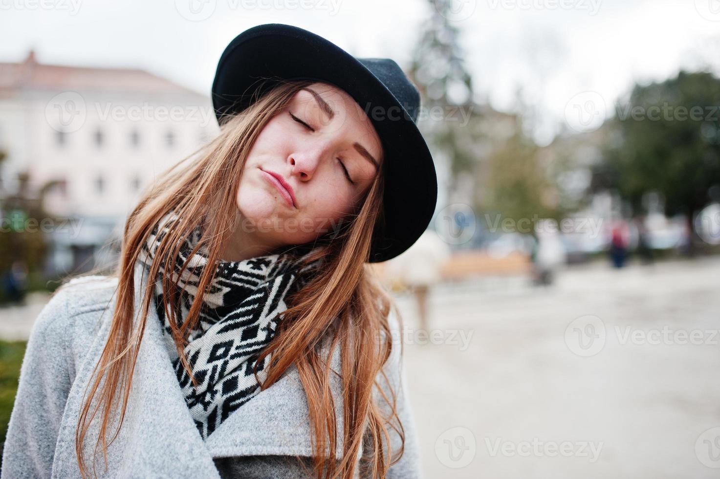 Cierra el retrato de una joven modelo divertida con un abrigo gris y un sombrero negro con los ojos cerrados y soñando en la calle de la ciudad. foto