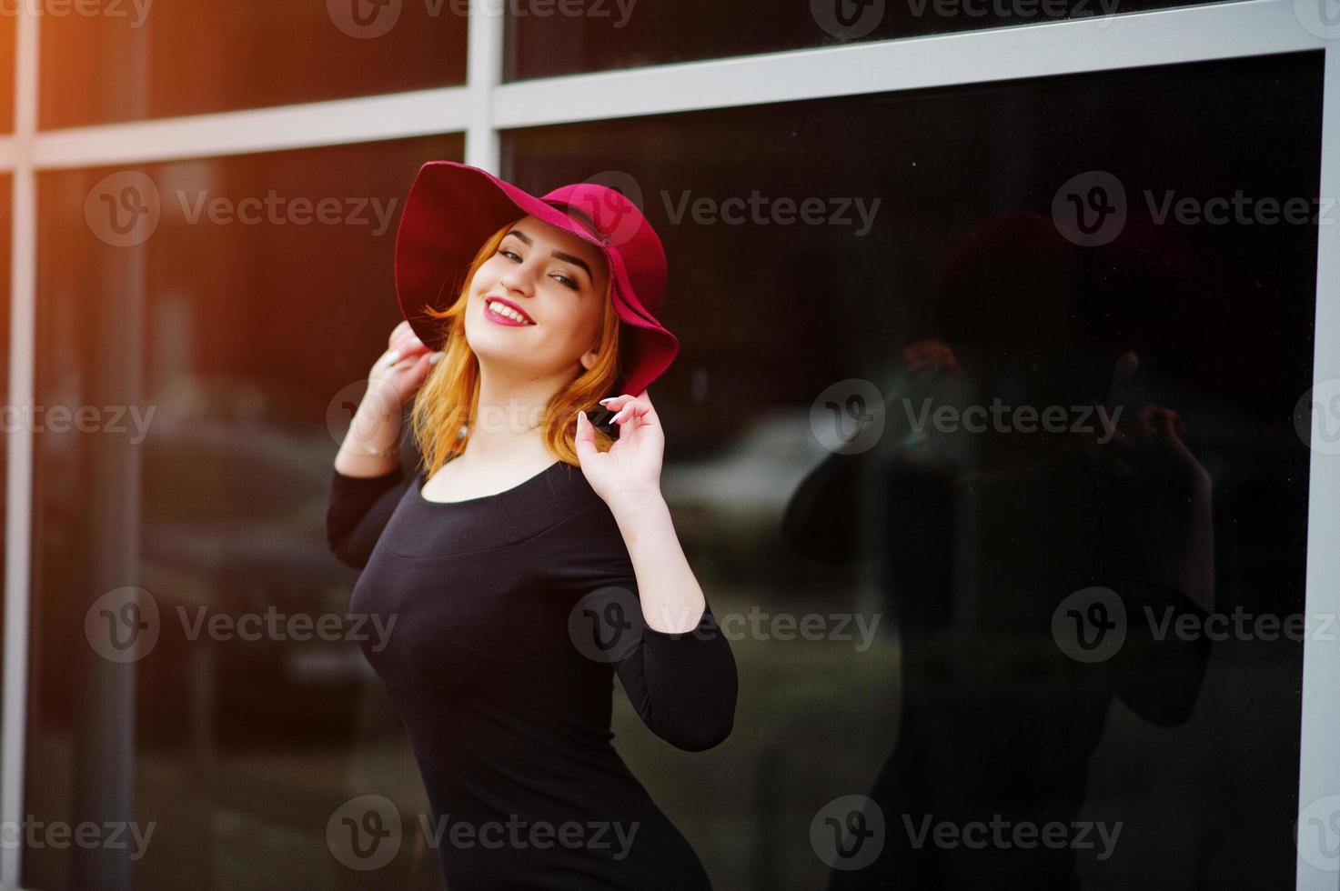 Portrait of fashion red haired girl on red hat and black dress with bright make up posed against large window. Photo toned style Instagram filters.