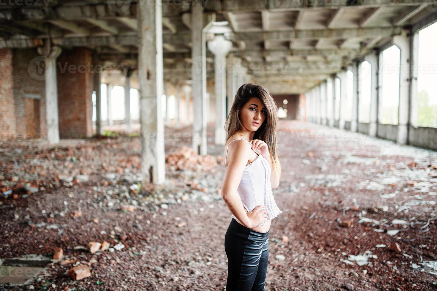 Portrait of young cute brunette girl wearing on black leather pants and white blouse posed on abandoned place. photo