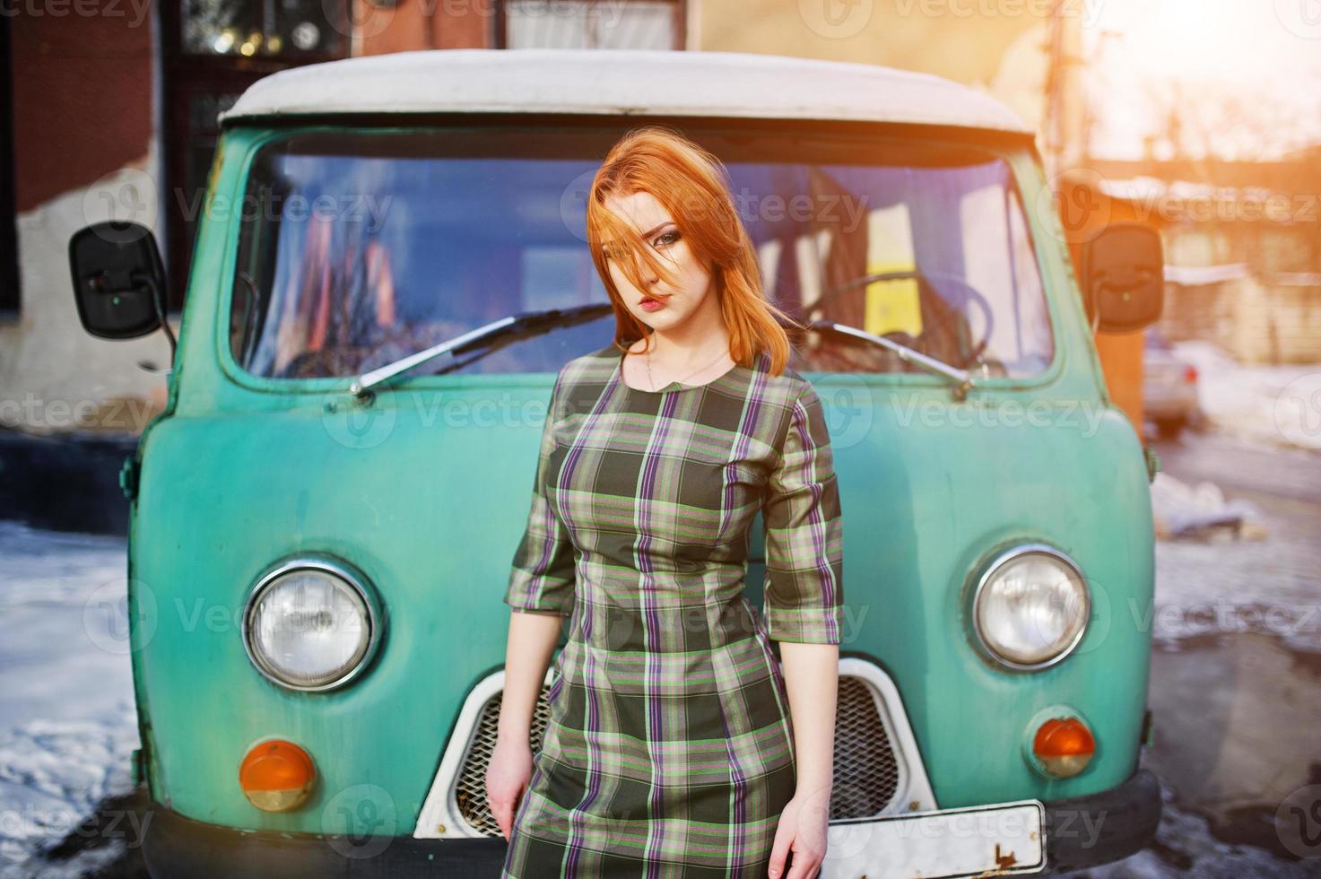 Young red haired girl posed on checkered dress background old retro cyan minivan. photo