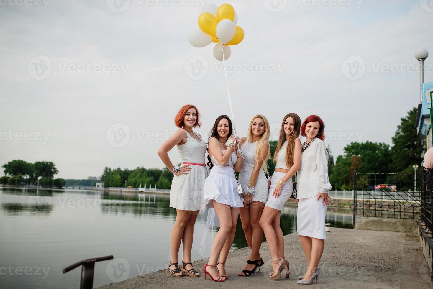 Five girls with balloons at hand weared on white dresses on hen party against pier on lake. photo