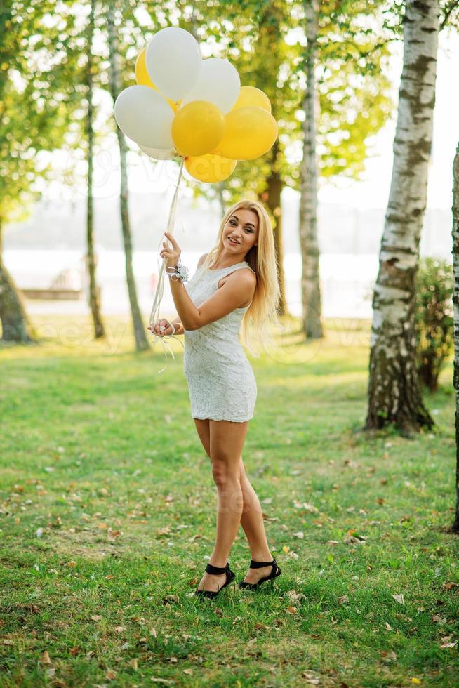 Portrait of blonde girl wearing on white dress with balloons at hands against park at hen party. photo