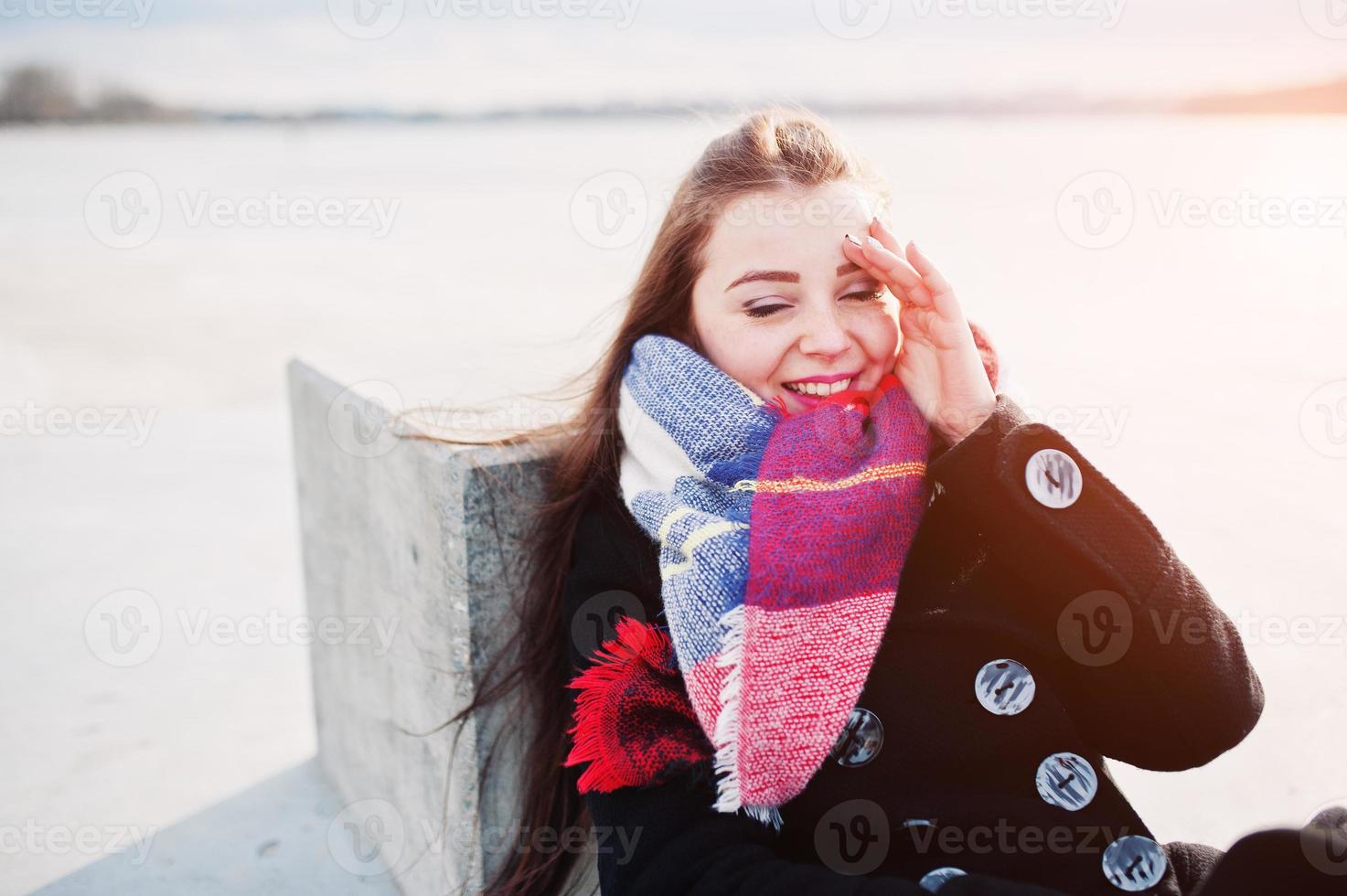 chica joven casual en abrigo negro, bufanda y sombrero contra el río congelado en el clima soleado de invierno. foto