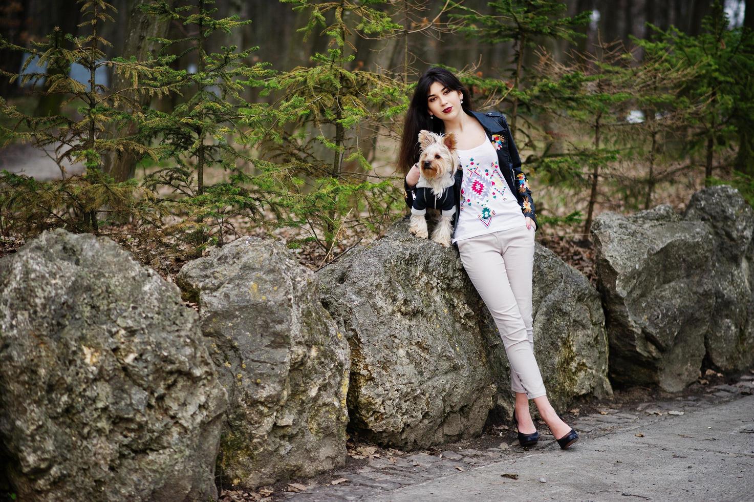 Brunette gypsy girl with yorkshire terrier dog posed against stones on park. Model wear on leather jacket and t-shirt with ornament, pants and shoes with high heels. photo