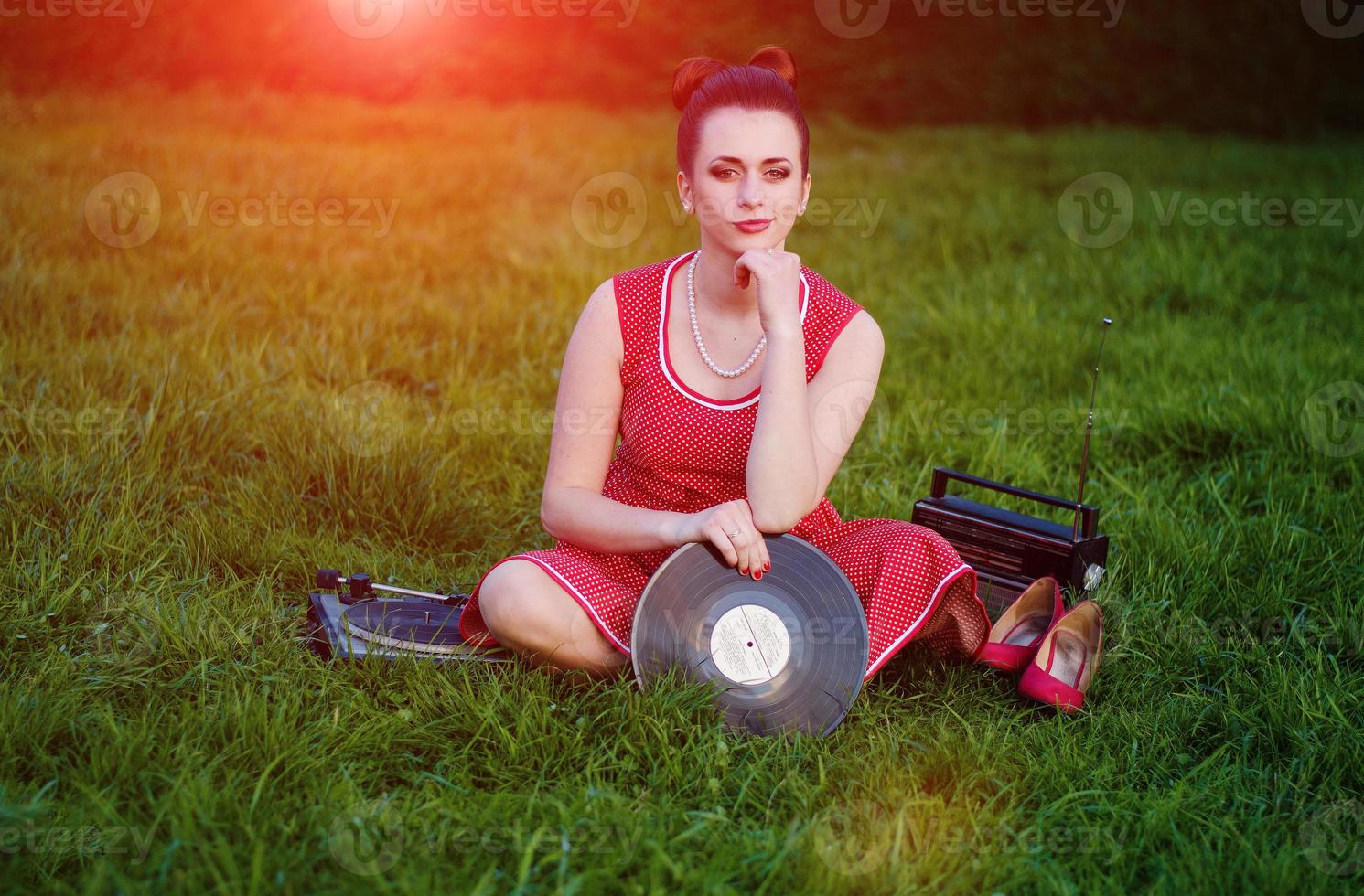 Portrait of young pinup girl wearing at retro vintage old-fashioned dress in peas sitting on grass wit retro radio and vinyl audio record in hands. photo