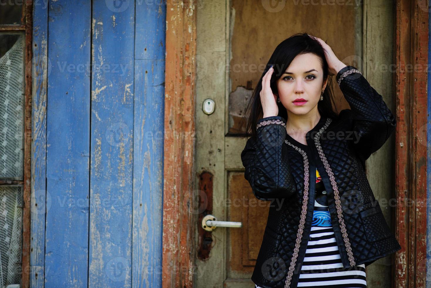 Brunette model girl at leather jacket posed background old wooden door of house. photo