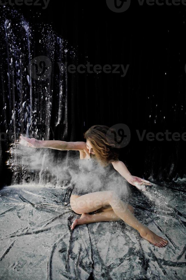 Girl dancer jumping and dancing in the white dust with flour on a black background. Studio shot of woman dancing with flour. photo