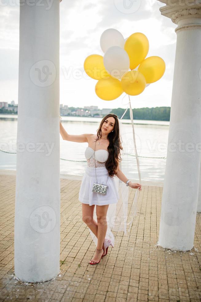 Young brunette girl with balloons at hand weared on white dress on hen party against white columns of arch. photo