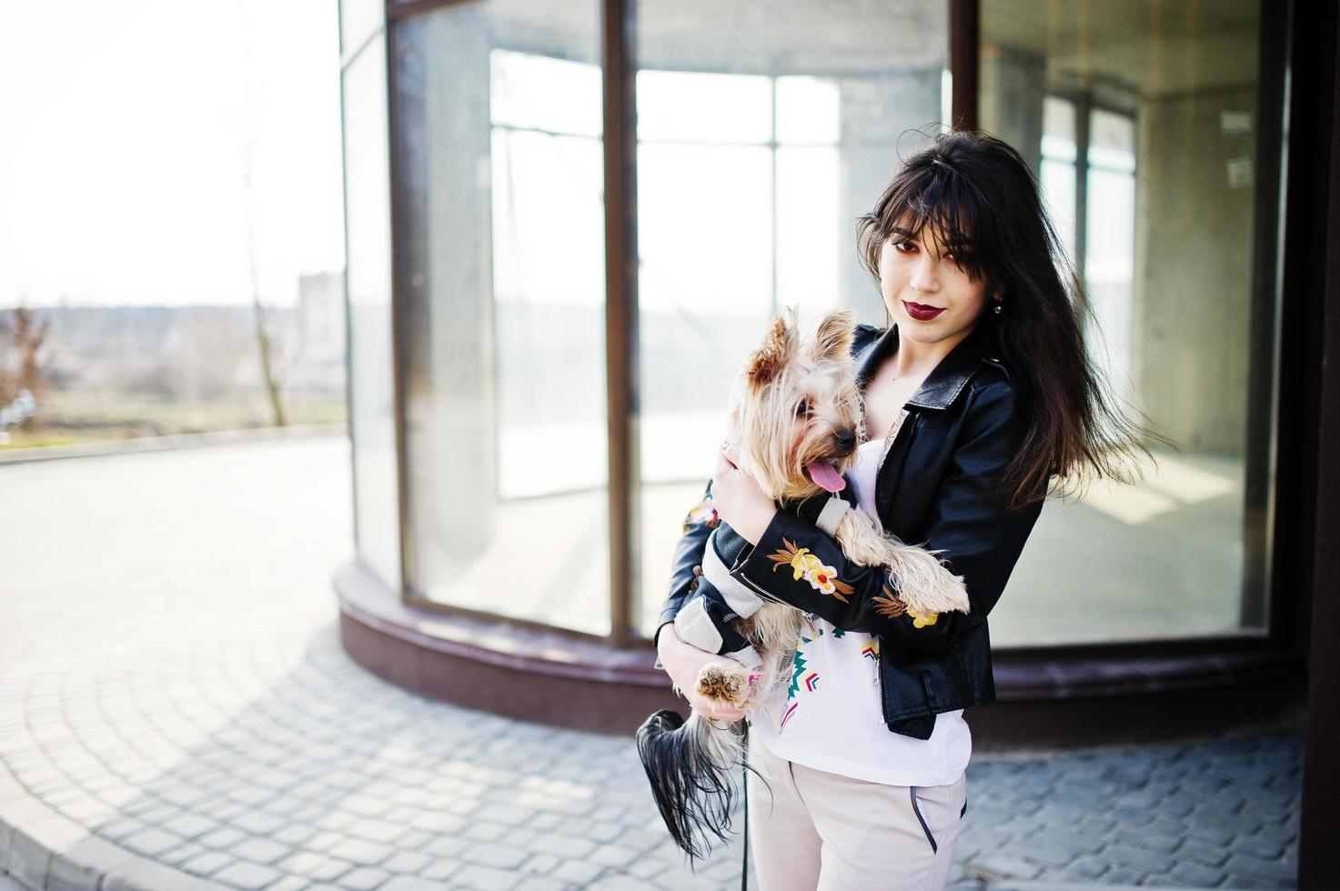 Brunette gypsy girl with yorkshire terrier dog posed against large windows house. Model wear on leather jacket and t-shirt with ornament, pants. photo