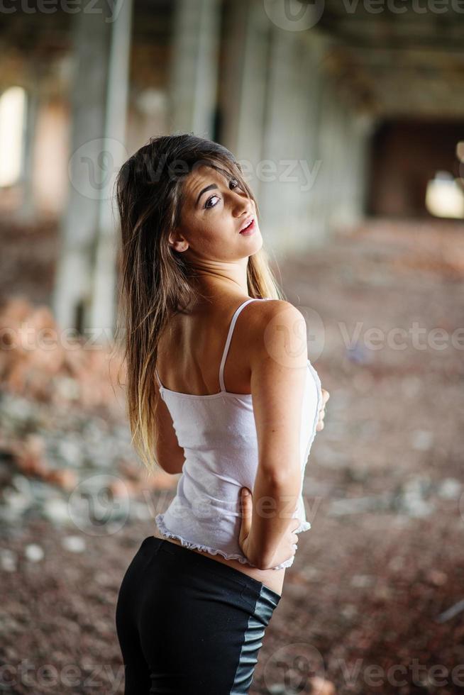 Portrait of young cute brunette girl wearing on black leather pants and white blouse posed on abandoned place. photo