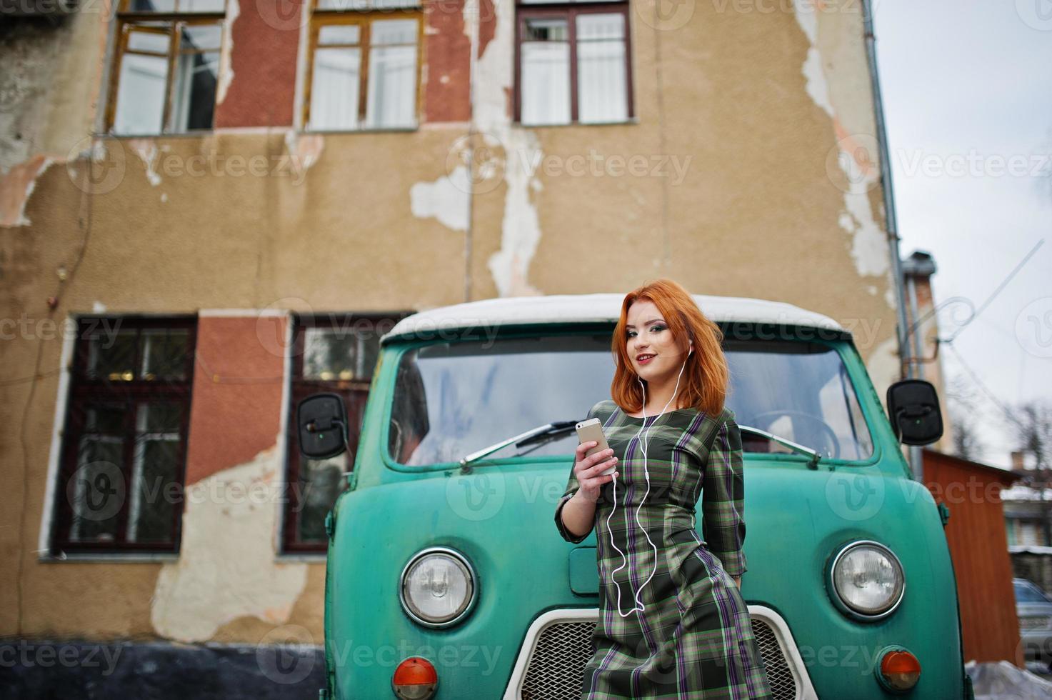 Young red haired girl lady with mobile cell phone and headphones, wearing on checked dress background old vintage turqoise minivan bus. photo