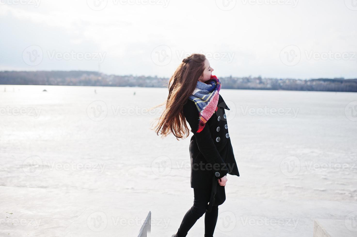 chica joven casual en abrigo negro, bufanda y sombrero contra el río congelado en el clima soleado de invierno. foto