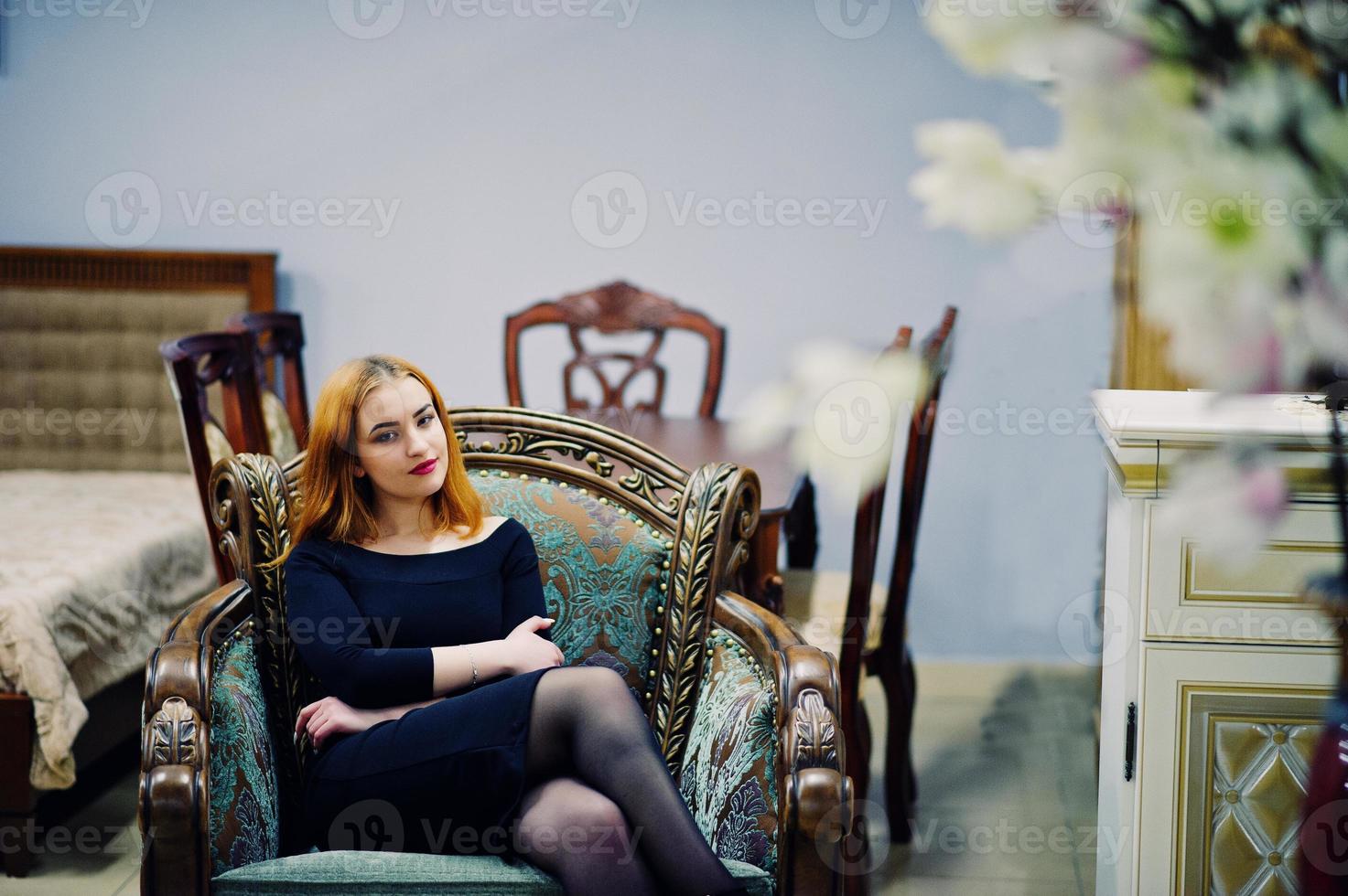 Fashion red haired girl wear on black dress with bright make up sitting on royal armchair. photo