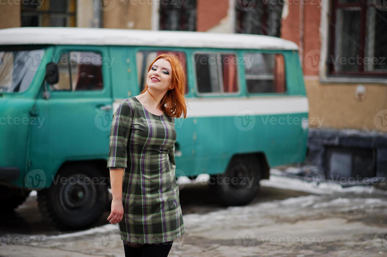 Young red haired girl posed on checkered dress background old retro cyan minivan. photo