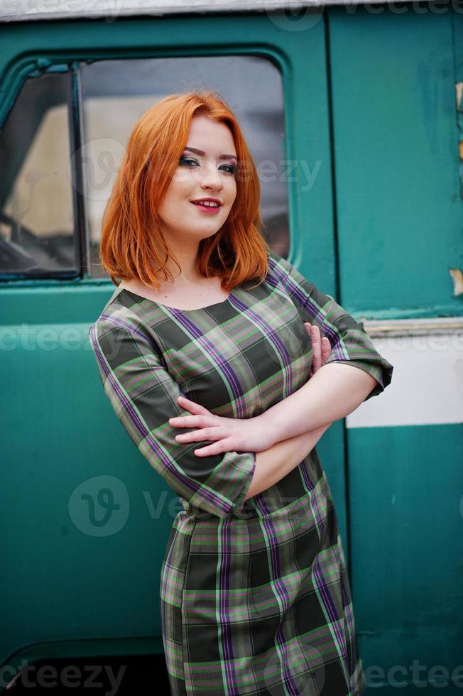 Young red haired girl posed on checkered dress background old retro cyan minivan. photo