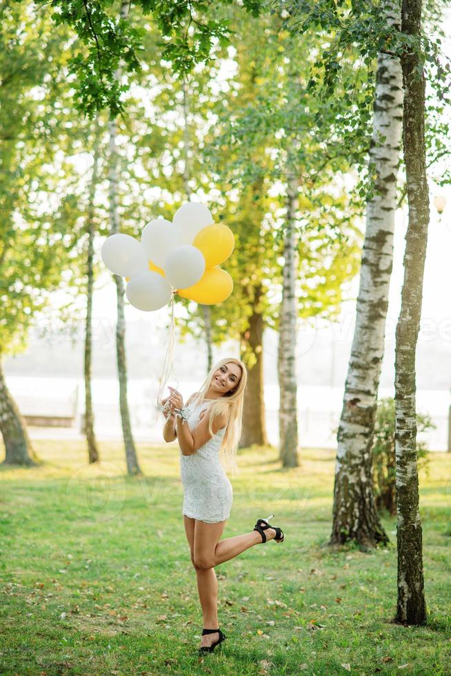 Portrait of blonde girl wearing on white dress with balloons at hands against park at hen party. photo