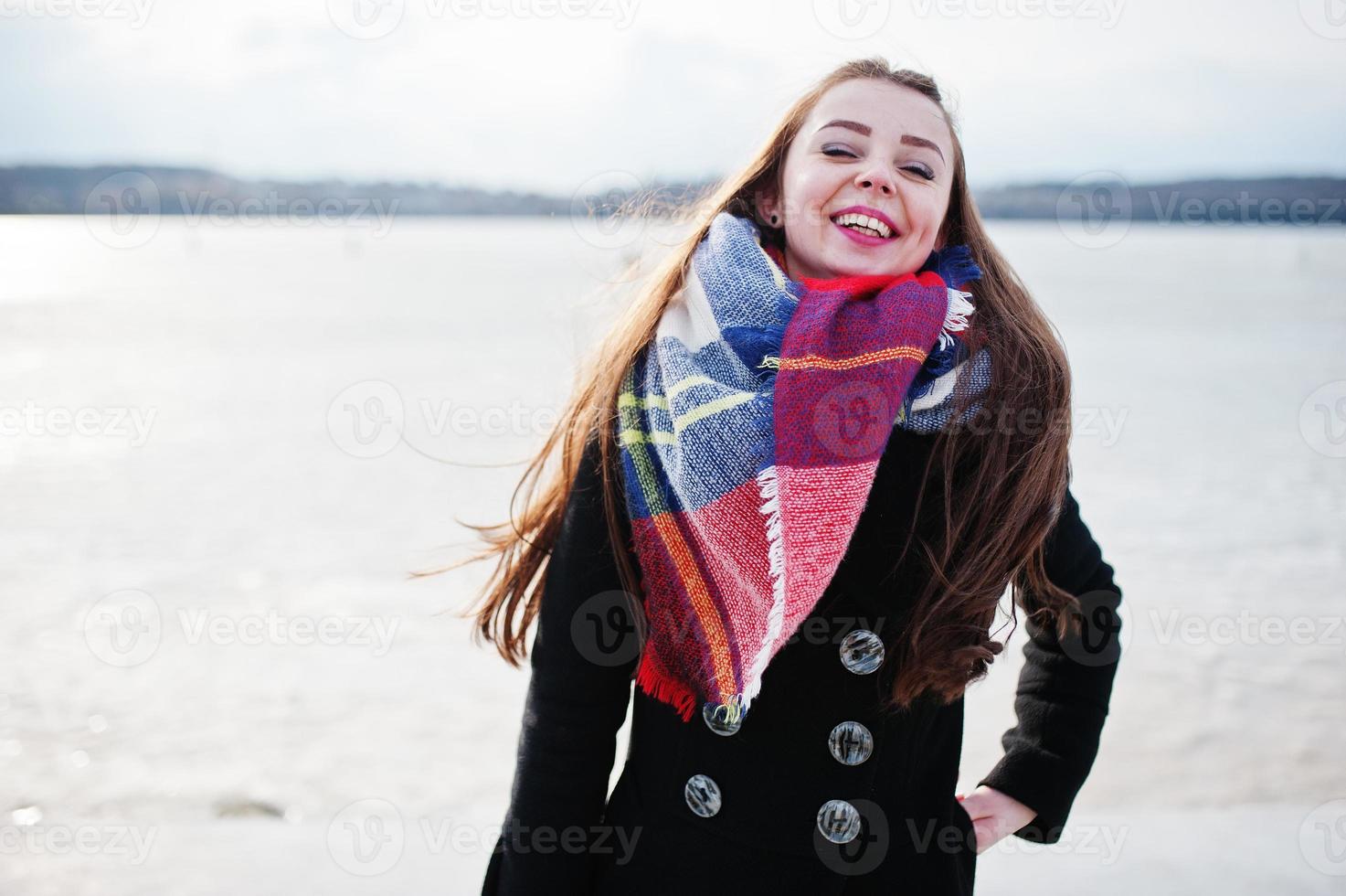 Casual young girl at black coat, scarf and hat against frozen river on sunny winter weather. photo