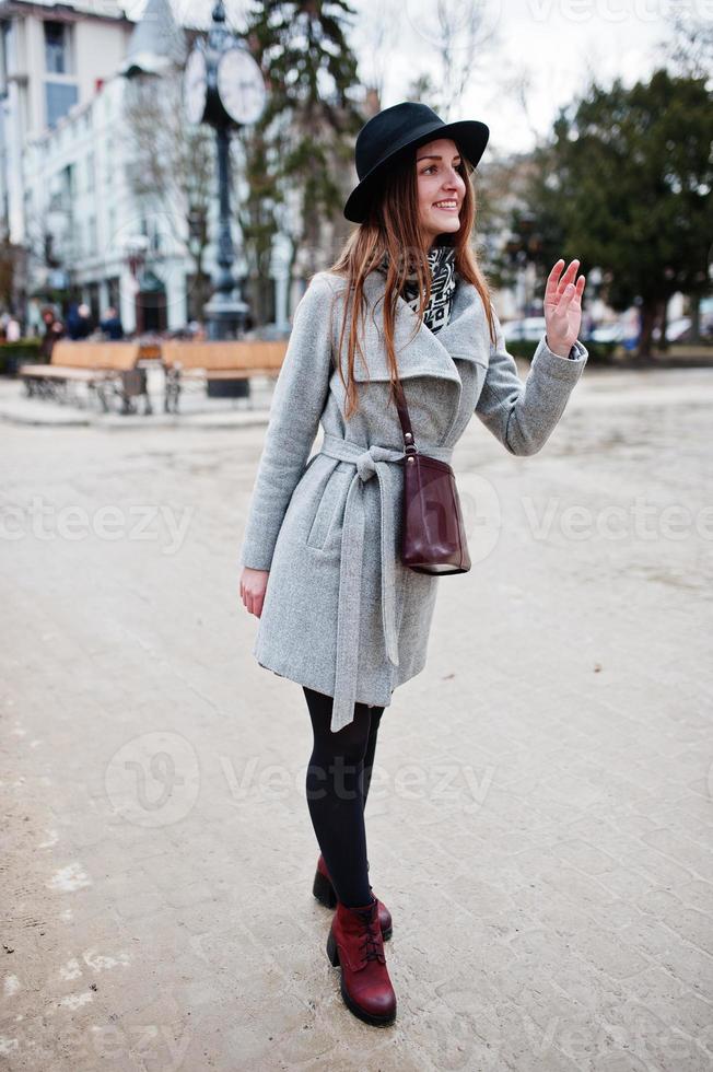 joven modelo turista con abrigo gris y sombrero negro con bolso de cuero sobre los hombros posada en la calle de la ciudad. foto