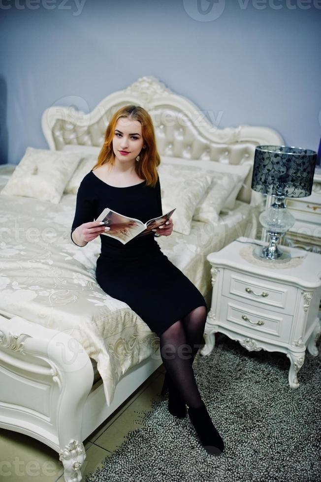 chica pelirroja con vestido negro sentada en la cama y leyendo una revista de moda. filtros de instagram de estilo foto tonificado.