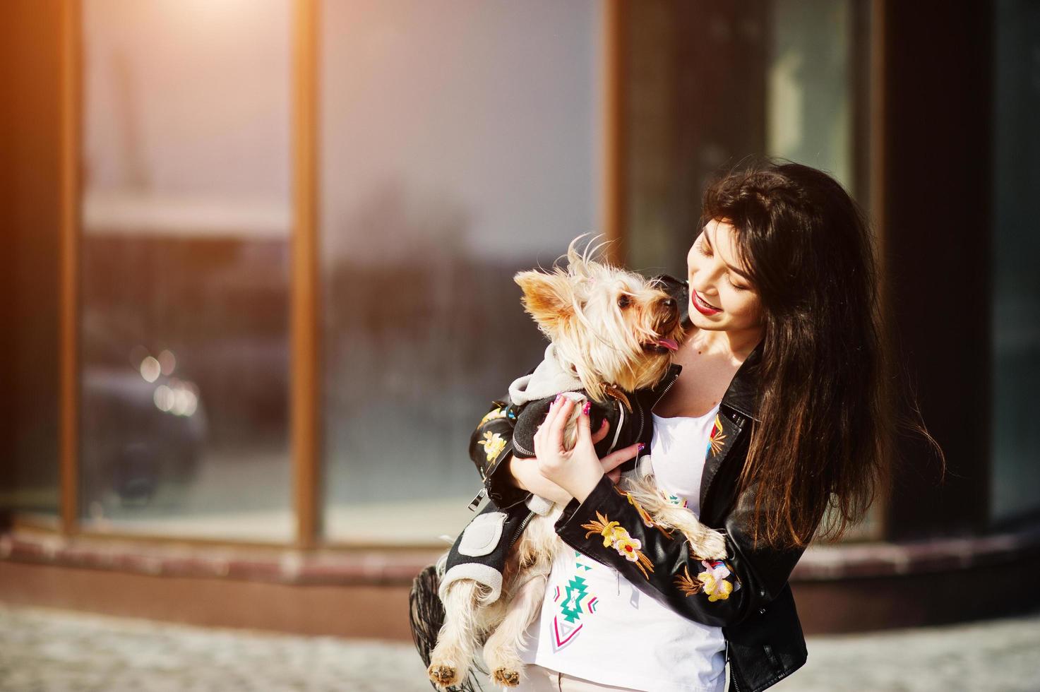 chica gitana morena con perro yorkshire terrier posó contra la casa de grandes ventanales. desgaste modelo en chaqueta de cuero y camiseta con adorno, pantalones. foto