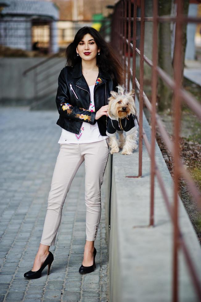 Brunette gypsy girl with yorkshire terrier dog posed against steel railings. Model wear on leather jacket and t-shirt with ornament, pants and shoes with high heels. photo
