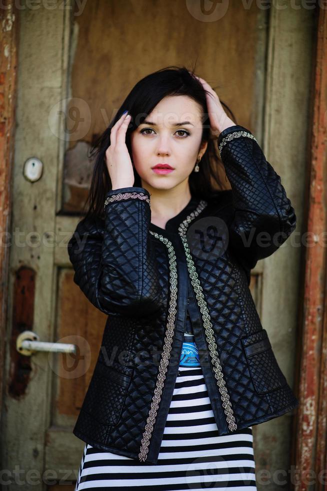 Brunette model girl at leather jacket posed background old wooden door of house. photo