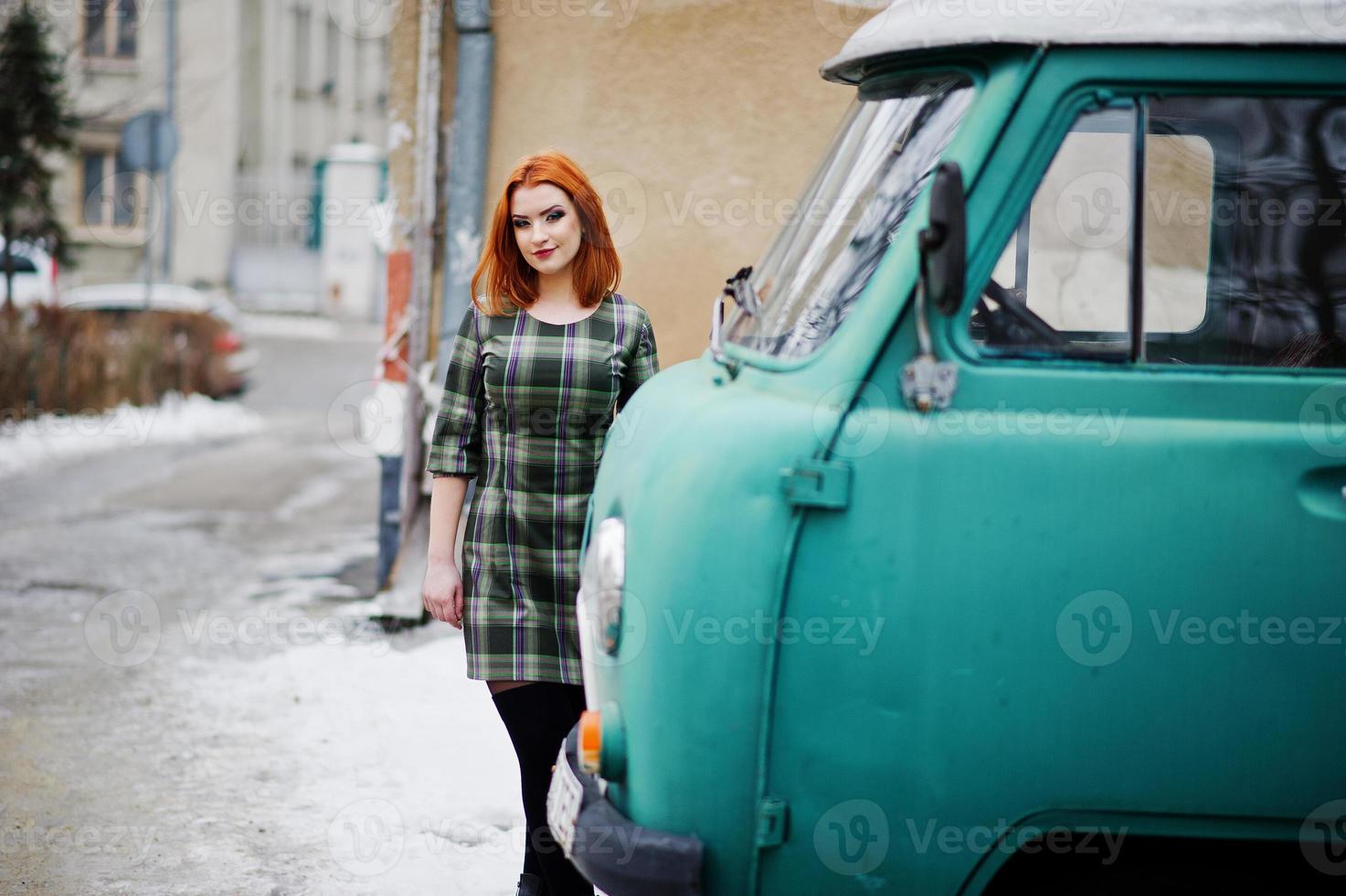 Young red haired girl posed on checkered dress background old retro cyan minivan. photo